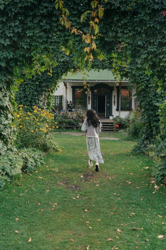 A woman runs through a lush green garden toward a charming house.