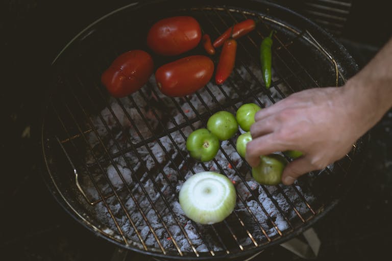 A hand grilling fresh vegetables like tomatoes, chilies, and onion over charcoal.