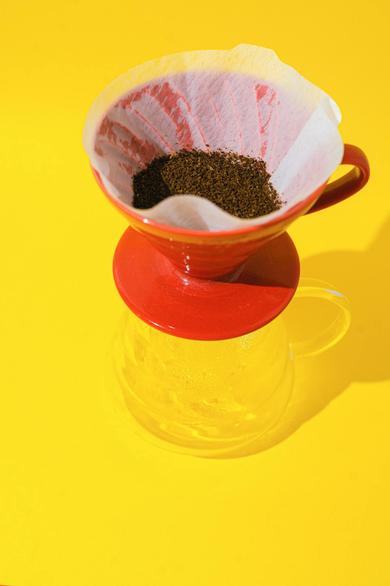 A vibrant close-up of drip coffee preparation using a red coffee dripper and filter paper against a yellow background.